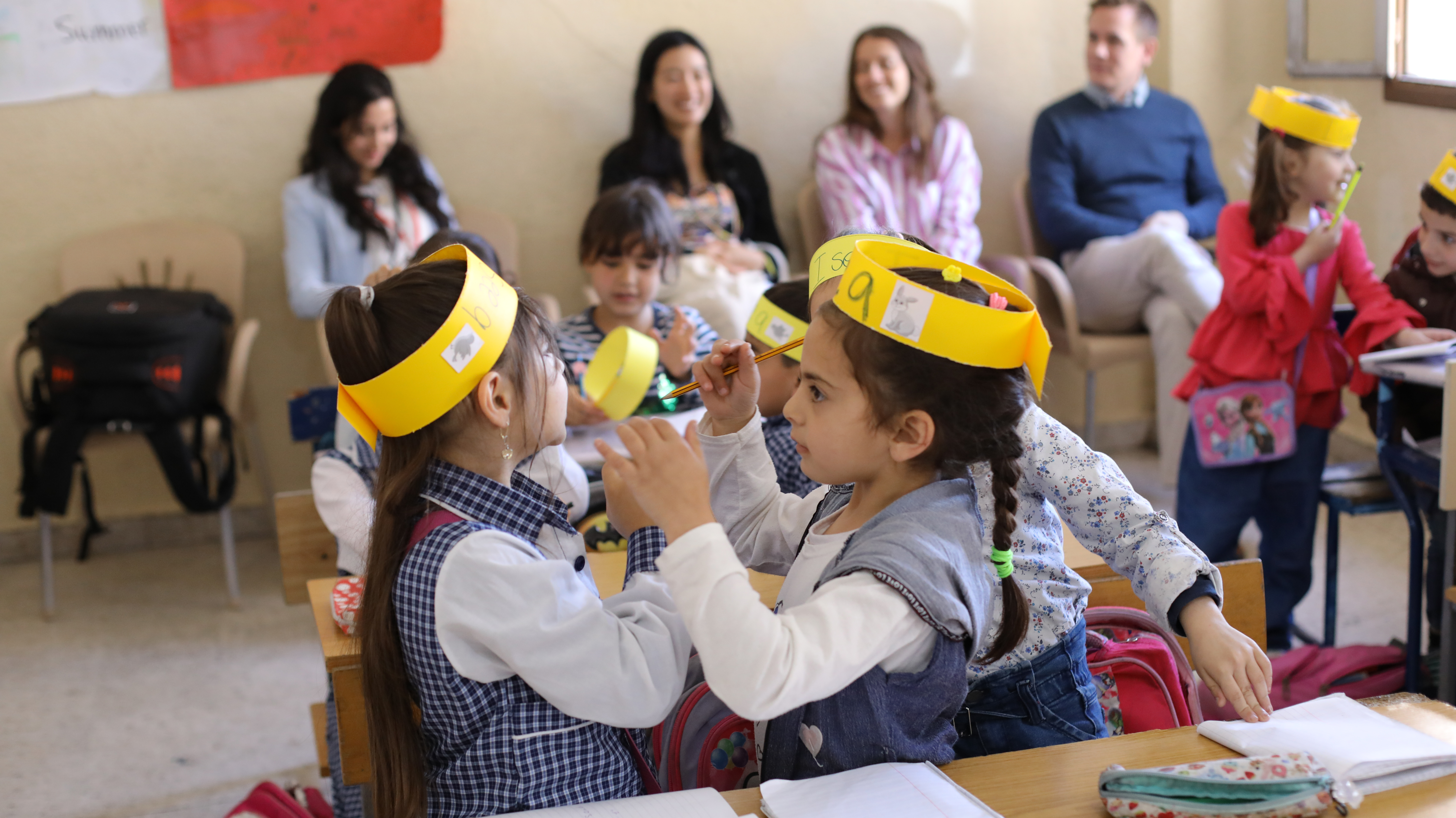Preview image for the video "Education in Emergencies: Connecting across borders" - Two young girls in a classroom wearing yellow paper headbands face each other.