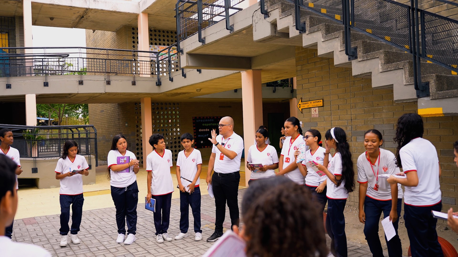 Preview image for the video "In Mauricio Bayona's classroom, everyone is a learner and a leader - Enseña por Colombia" - A young male teacher in a white shirt with a shaved head stands in a courtyard speaking, surrounded by teenage students also in white shirts.