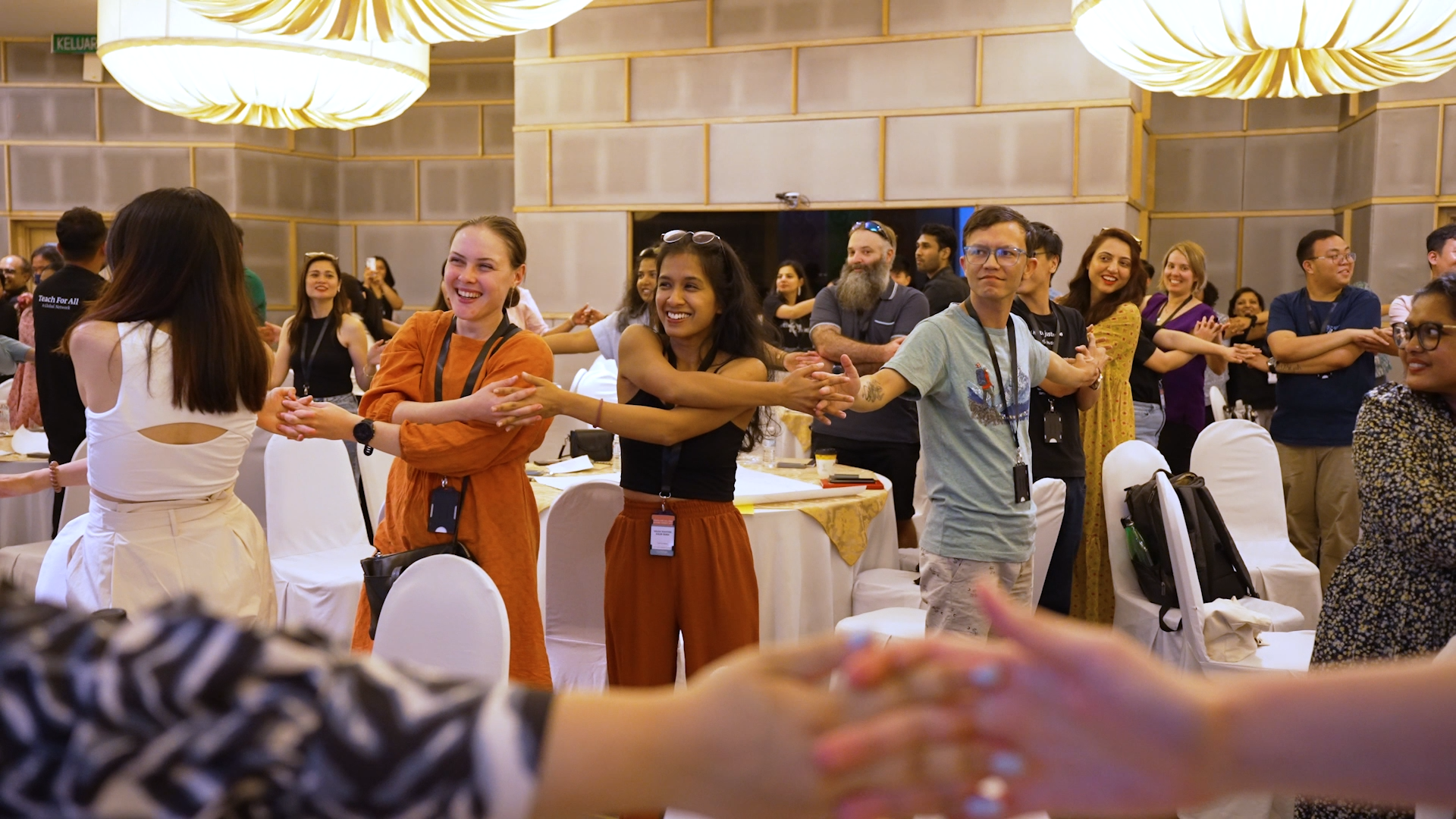 Preview image for the video "Alumni Impact Across the Teach For All Network in Asia Pacific" - A large group of young adults hold hands while standing in a long chain in a conference room.