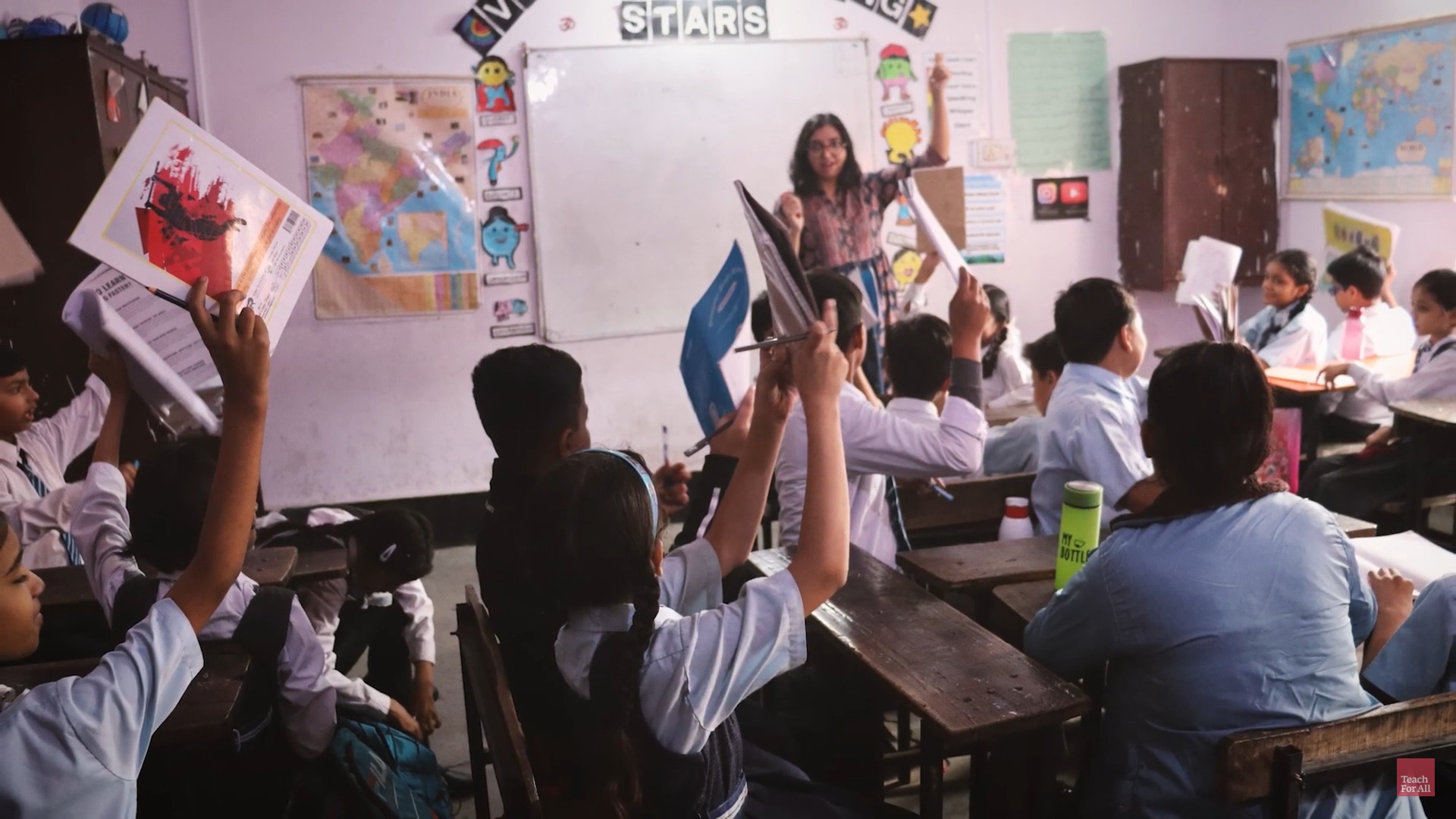 Preview image for the video "Richa Gupta - Teach For India" - A classroom full of young Indian chilren raise notebooks and smile at their young female teacher standing in the front of a whiteboard.