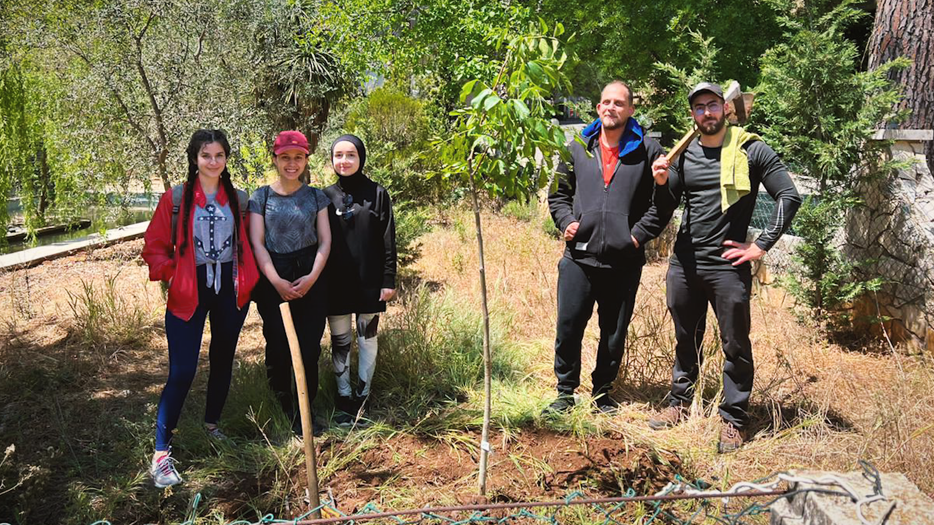 Preview image for the video "Ahmad El Baghdadi - Teach For Lebanon" - Two bearded men holding shovels and three young teen girls stand near two freshly planted saplings.