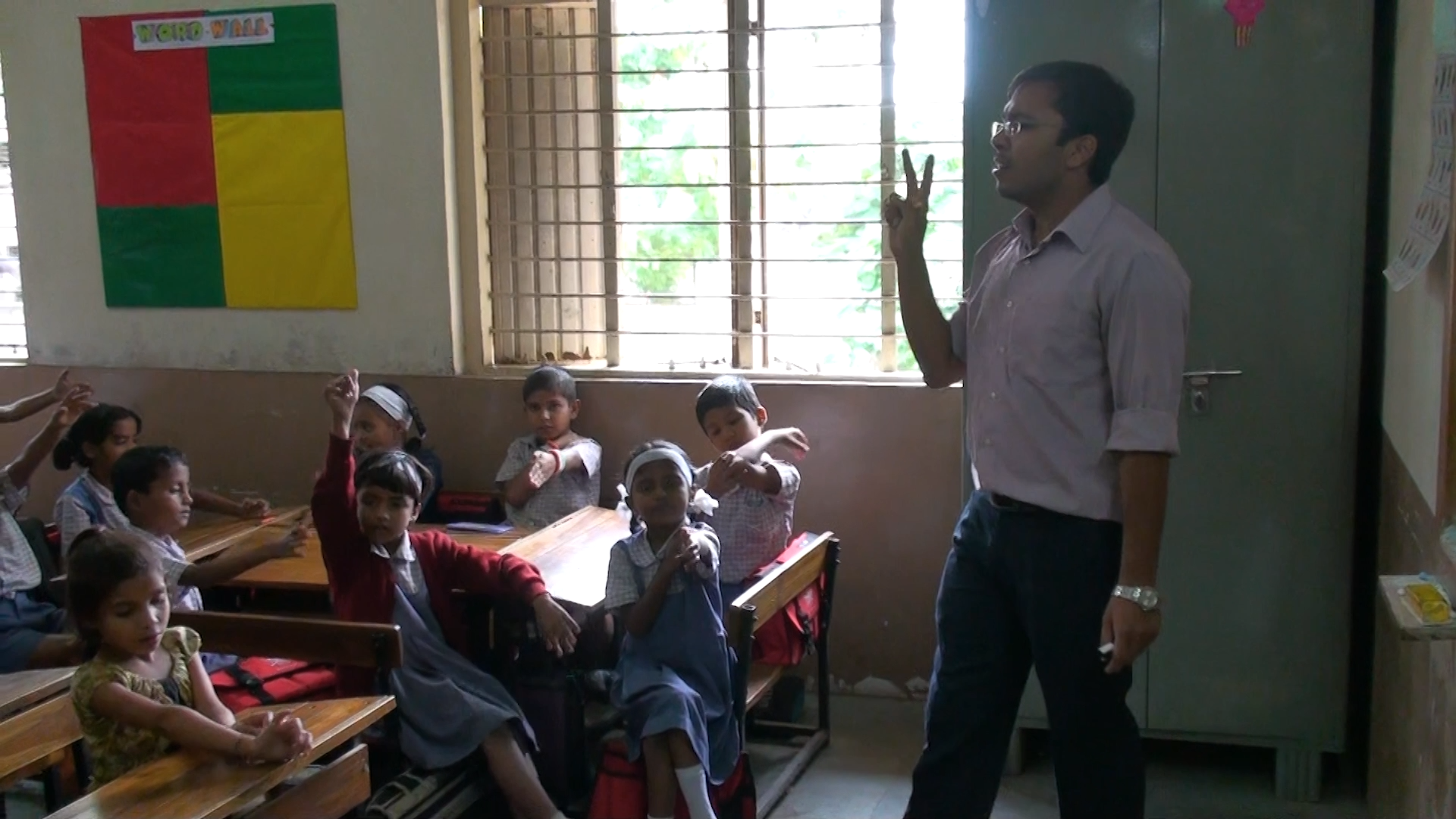 Preview image for the video "Mayank Lodha -  Teach For India" - A young Indian male teacher stands in front of his class of young students holding up two fingers.