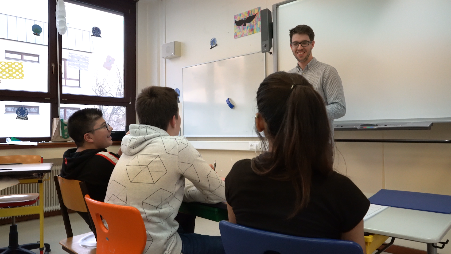 Preview image for the video "Felix Stadler: Teach For Austria alum, teacher &amp; politician for equitable education." - Through the backs of students' heads we see a young white male teacher with brown hair and glasses standing in front of a whiteboard..