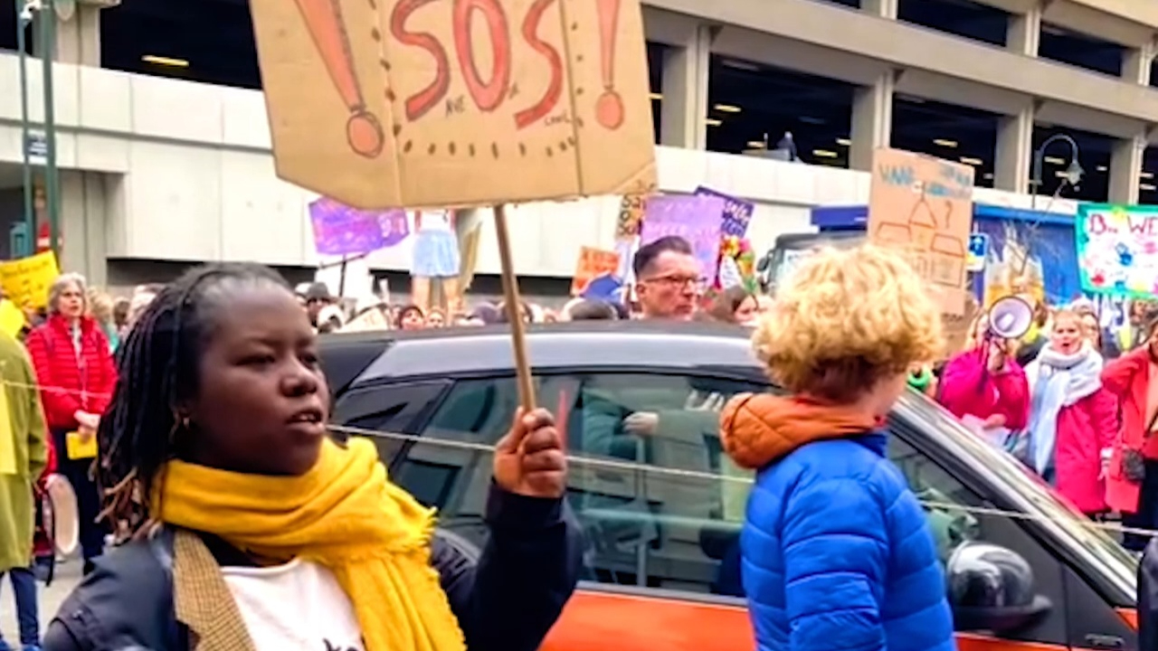 Preview image for the video "In Belgium, Ange Raïssa Uzanziga is Helping Students Overcome Barriers to Education and Thrive" - A Black woman with a yellow scarf around her neck holds a sign that says SOS in what appears to be a protest in the city streets.