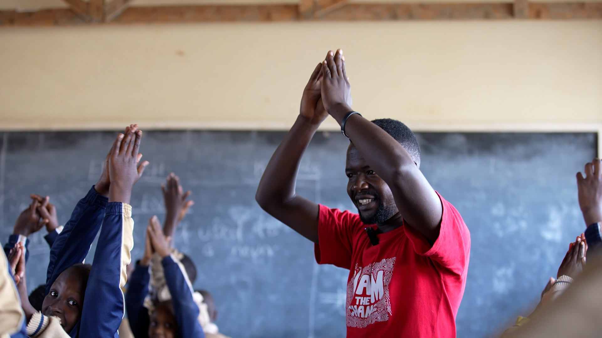 Preview image for the video "How a Teacher in Kenya Turned His Struggle Into Student Confidence" - A young Black man claps his hands over his head in a classroom with students doing a similar gesture in front of him.