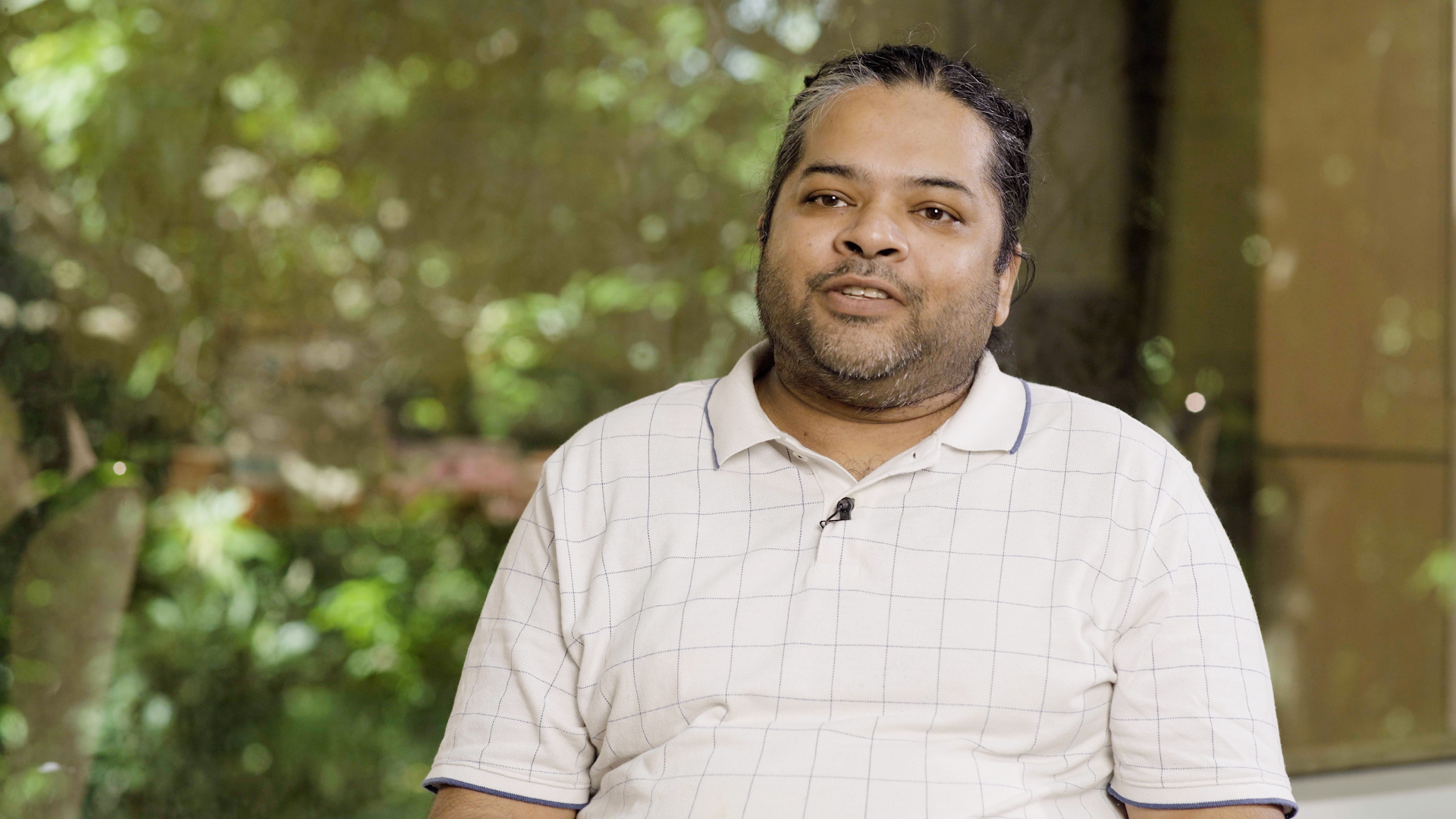 A medium shot of a man with tan skin and dark, salt-and-pepper hair pulled back, featuring a short beard and mustache. He is wearing a white short-slee polo shirt with a subtle grey grid pattern and a small black lapel microphone clipped to the collar. He is positioned in the center of the frame against a soft, out-of-focus background of lush green foliage and dappled sunlight.