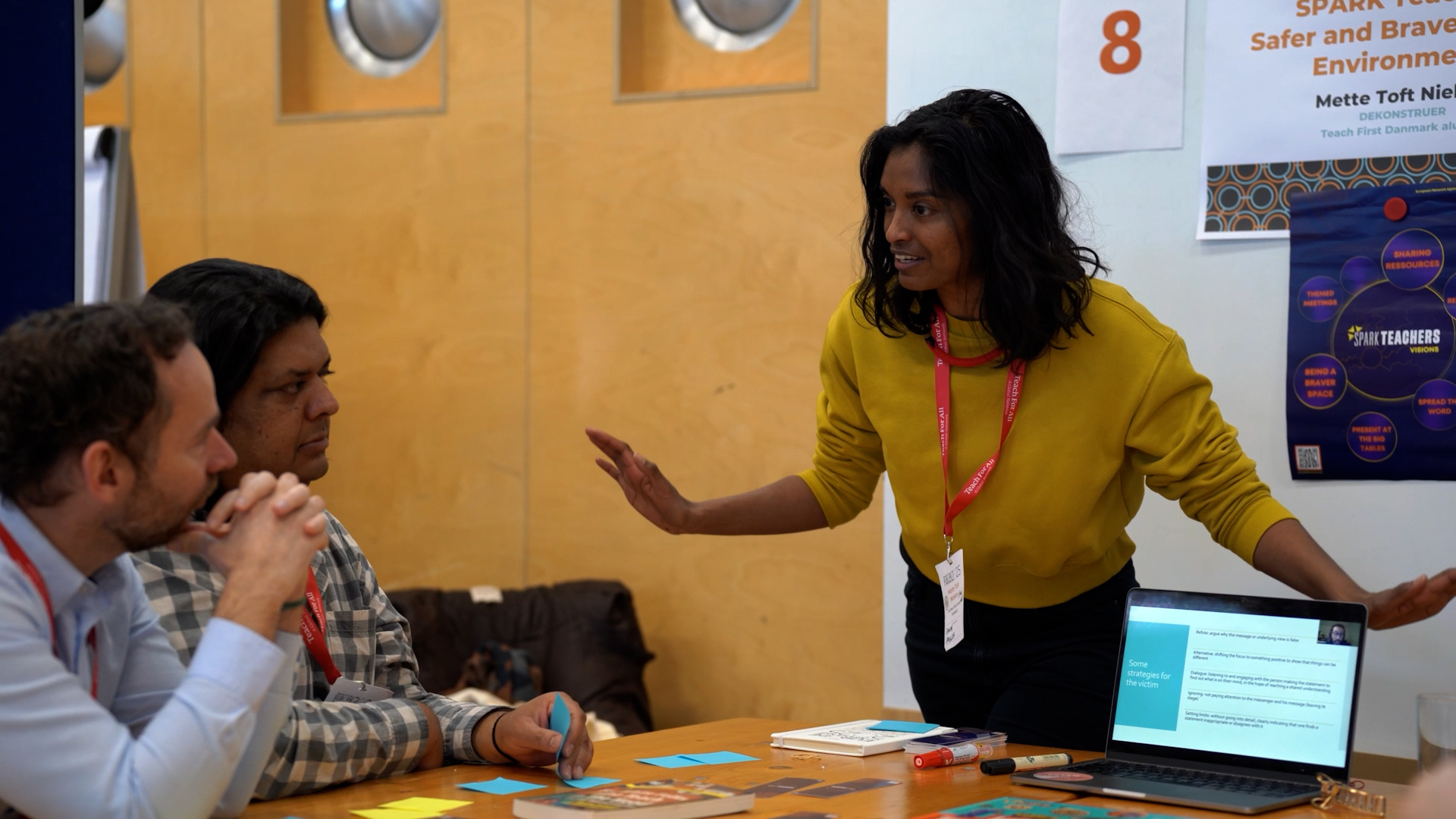A woman with brown hair and brown skin in a yellow sweatshirt gestures with both hands as she speaks to two men seated at a table during a workshop presentation.