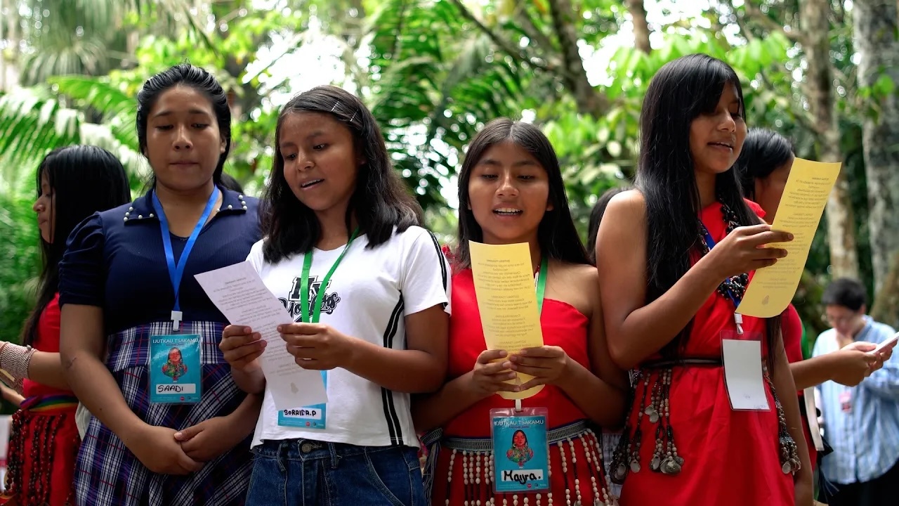 A group of young Indigenous women stand together outdoors surrounded by lush greenery, holding printed papers as they sing or read aloud during an event.