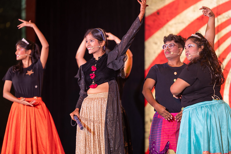 A group of five young performers in colorful costumes dance on a stage. In the center, a smiling young woman wearing a long black ruffled top and a sparkly gold skirt raises her left arm high. To her left, a dancer in a black t-shirt and a long bright orange skirt strikes a pose, while to her right, two performers in black shirts look on—one wearing a blue skirt and the other wearing pink patterned trousers and round white glasses.