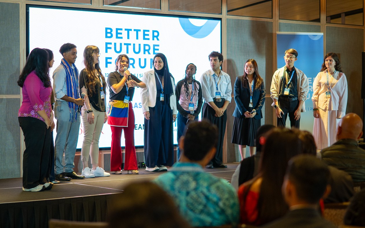 A group of ten young people stands on a stage facing an audience during a presentation. They are lined up in front of a large digital screen displaying the words "BETTER FUTURE FORUM." One young woman near the center-left holds a microphone and speaks, with an Ecuadorian flag tied around her waist. The other presenters look toward her or the audience, all wearing business casual attire, some with event lanyards. The back of the audience members' heads are visible in the foreground.