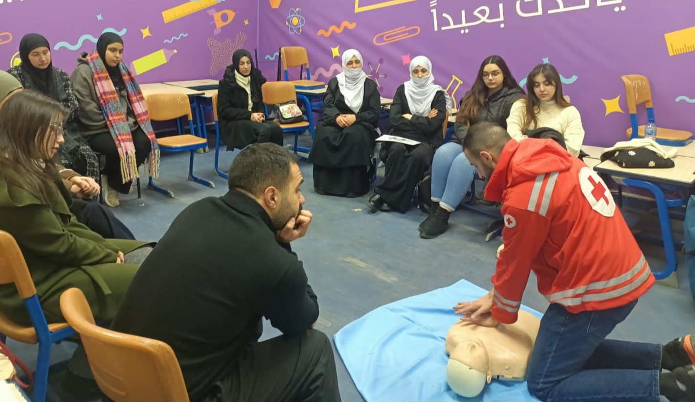 A man in a Red Cross jacket demonstrates CPR to men and women seated in a circle, some of the women are wearing head scarves