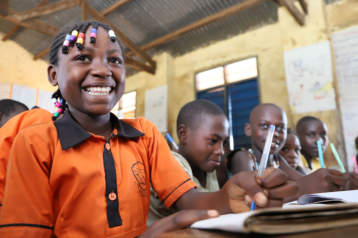 A close-up, low-angle shot of a young Black schoolgirl smiling brightly at her desk in a classroom. She has braided hair with vibrant pink, yellow, and blue beads, and wears an orange polo uniform with a school crest. She is holding a blue pen over a notebook. In the background, other young classmates sit at wooden desks writing, inside a classroom with yellow walls and a visible corrugated metal roof structure.