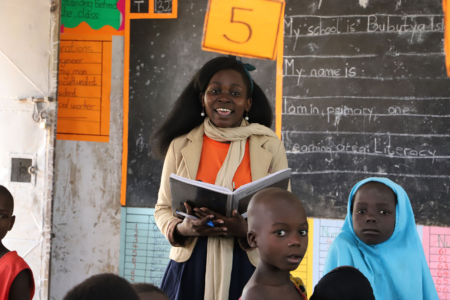 A smiling woman stands in front of a classroom blackboard holding an open notebook, addressing a group of young students in the foreground. She wears a beige jacket over an orange t-shirt and a light-colored scarf. The blackboard behind her has sentence frames written in chalk, including "My school is" and "My name is," alongside a bright orange sign displaying the number "5."