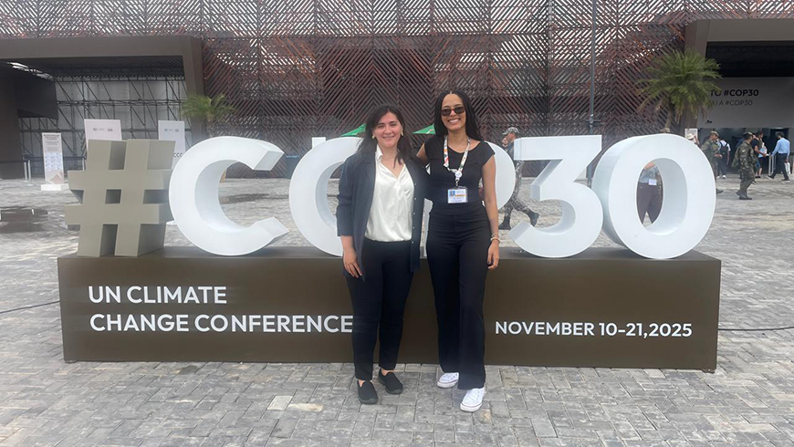 two women in business clothin stand in front of a large COP30 sign