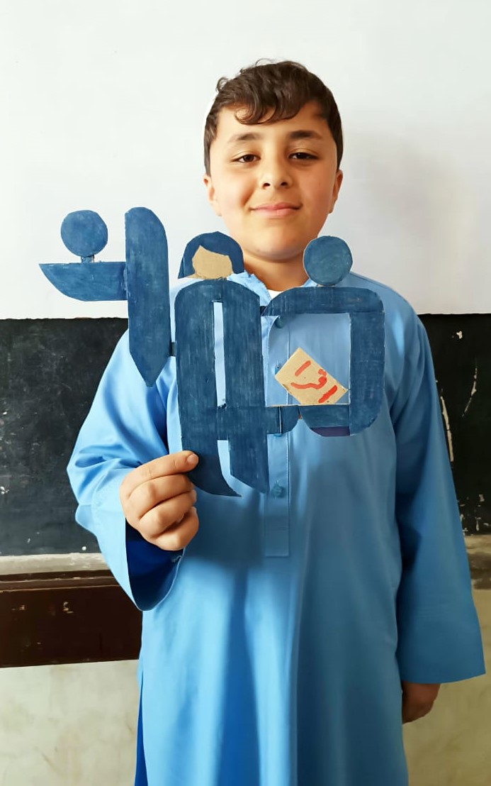 A smiling young boy in a light blue tunic holds up a handmade blue cardboard cutout of the Teach For Afghanistan (TAO) logo.
