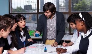 A light skinned young man with brown hair and a beard demostrates a science experiment using beekers to a diverse group of teenagers