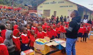 A young Black man stands in front of a bench piled with workbooks and a large crowd of Black children in school uniforms outside of a school