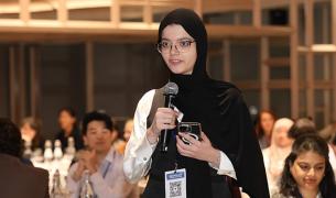 A young woman wearing a black head scarf and glasses speaks into a microphone 