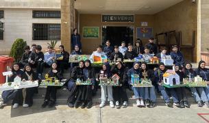 Many children in school uniform sit on steps outside their schools holding models of homes