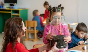  A young girl with braided hair and yellow clips holds a large glass jar containing soil and small green sprouts. An adult woman in a red sweater sits beside her, steadying the bottom of the jar as they look at the plants together. In the background, other children are seated at tables in a brightly lit classroom, including a boy to the right using a magnifying glass.