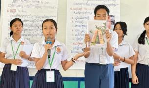 A group of school students in white short-sleeved shirts and dark skirts or trousers stand in front of a white board giving a presentation. A young woman in the front left holds a microphone and speaks, gesturing with her left hand toward a classmate beside her. The young man holds up a printed photograph showing two decorated bamboo bottles nestled in green leaves. In the background, posters with text in English and Khmer script are visible, with one heading reading "Topic: Bamboo bottles for our community