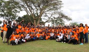 A wide shot of a large group of people posing together on a grassy lawn in front of a sprawling, leafy tree under an overcast sky. The majority of the group is wearing matching bright orange t-shirts with a graphic logo on the front, while a few individuals are in business or white shirts. Many people in the crowd are smiling and waving their hands at the camera. The individuals are arranged in rows, with those in the front kneeling or sitting on the grass, and those in the back standing.