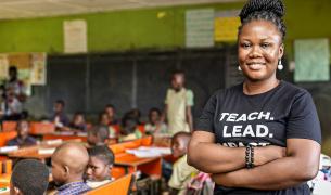 A smiling Black woman with her arms crossed wears a black T-shirt that reads "TEACH. LEAD." while standing in the foreground of a classroom filled with seated students.