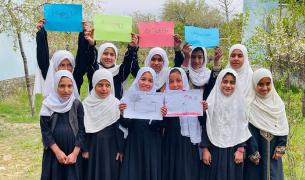 A group of young girls wearing white hijabs stand outdoors, holding up colorful signs that read "Happy 10th Anniversary Of Teach For Afghanistan."