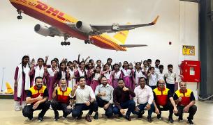 A group of school children in uniforms and several adults pose together indoors, with a large model of a DHL cargo plane mounted on the wall above them.