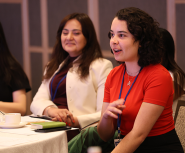 A young woman with shoulder-length brown curly hair and pale skin wearing a red shirt speaks while seated as others at the table watch and listen