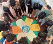 A group of children form a circle with their arms around each other looking down at a paper flower in the middle of the circle