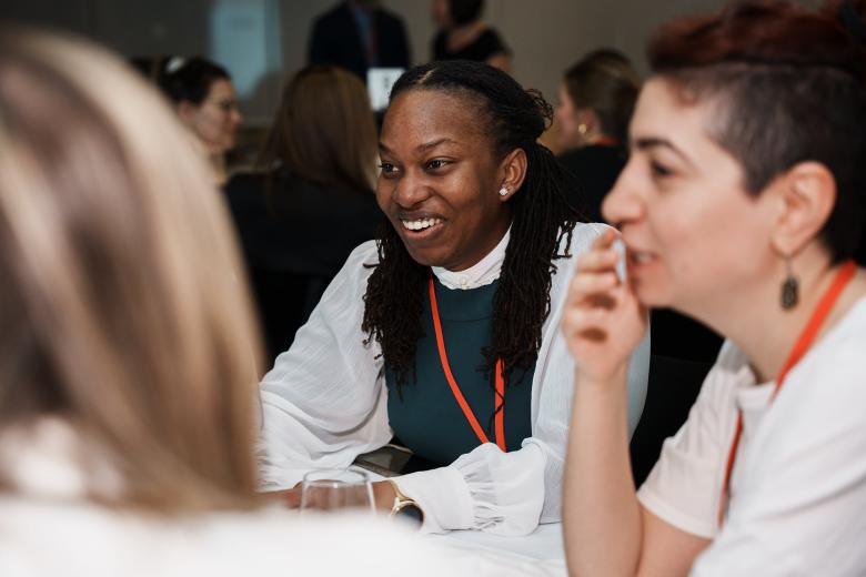 A young Black woman smiling sits at a table next to a white soman with short brown hair, both wearing red lanyards