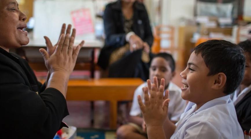 In a classroom, a young boy with brown hair and an older woman in a black sweater look at each other while clapping their hands