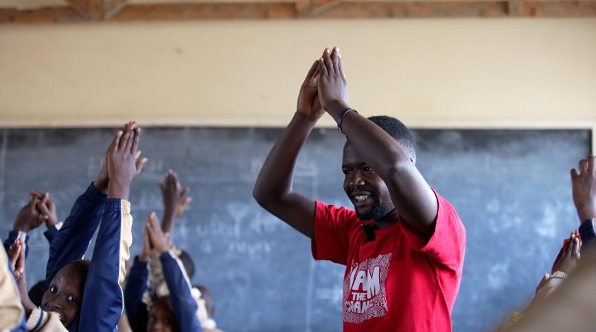 A young Black man claps his hands over his head in a classroom with students doing a similar gesture in front of him