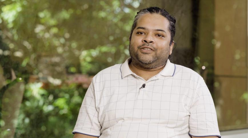 A medium shot of a man with tan skin and dark, salt-and-pepper hair pulled back, featuring a short beard and mustache. He is wearing a white short-slee polo shirt with a subtle grey grid pattern and a small black lapel microphone clipped to the collar. He is positioned in the center of the frame against a soft, out-of-focus background of lush green foliage and dappled sunlight.