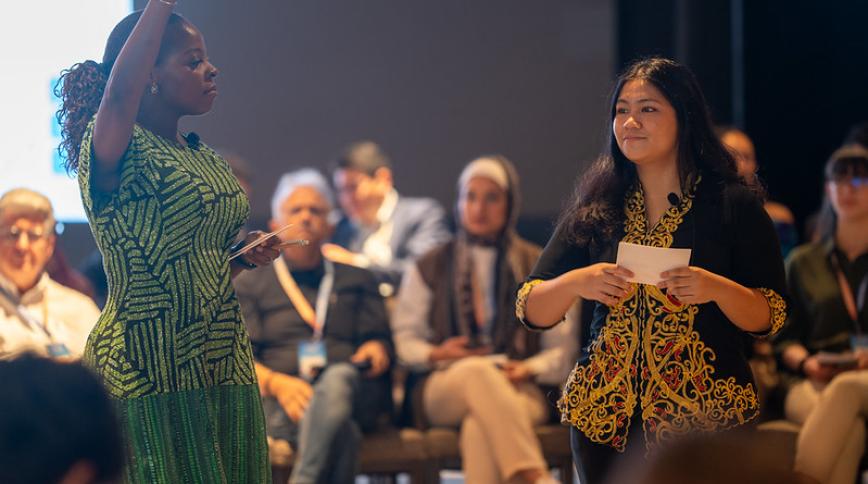 Two women present to an audience seated in a semi-circle. On the left, a Black woman in a patterned green dress raises her right hand while holding notes. On the right, a East Asian woman smiles while holding a note card, wearing a black top decorated with intricate gold embroidery along the front and sleeves. Both wear small lapel microphones.