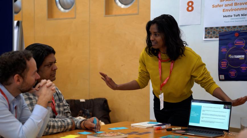 A woman with brown hair and brown skin in a yellow sweatshirt gestures with both hands as she speaks to two men seated at a table during a workshop presentation.