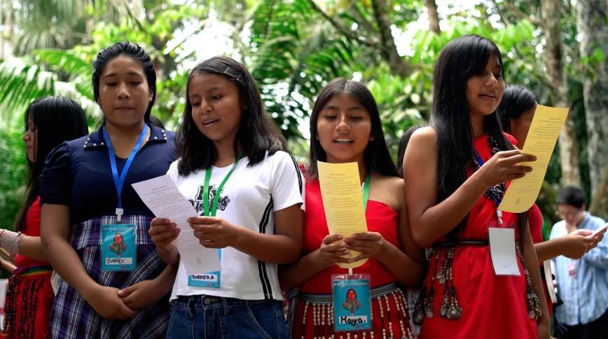 A group of young Indigenous women stand together outdoors surrounded by lush greenery, holding printed papers as they sing or read aloud during an event.