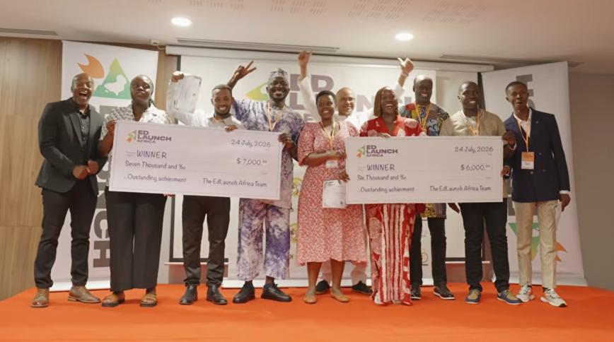 A group of nine Black men and women, some in cultural attire, pose and celebrate on stage as winners of EdLaunch Africa, holding two oversized presentation checks dated July 24, 2026.