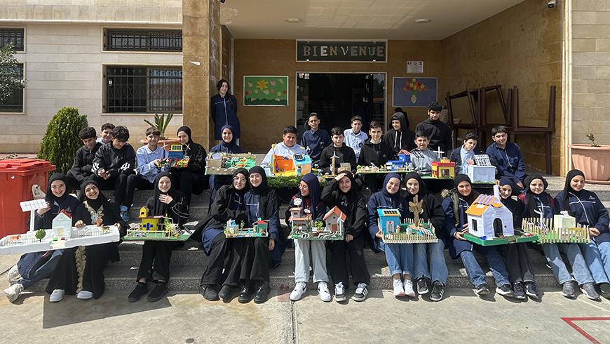 Many children in school uniform sit on steps outside their schools holding models of homes