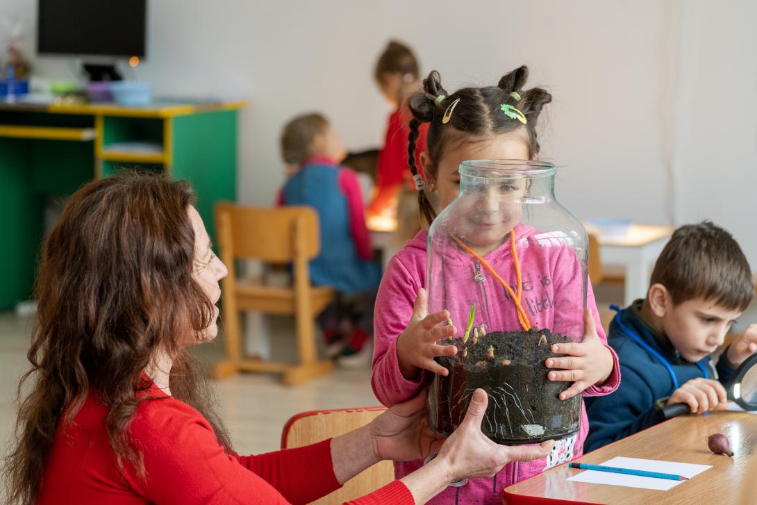  A young girl with braided hair and yellow clips holds a large glass jar containing soil and small green sprouts. An adult woman in a red sweater sits beside her, steadying the bottom of the jar as they look at the plants together. In the background, other children are seated at tables in a brightly lit classroom, including a boy to the right using a magnifying glass.