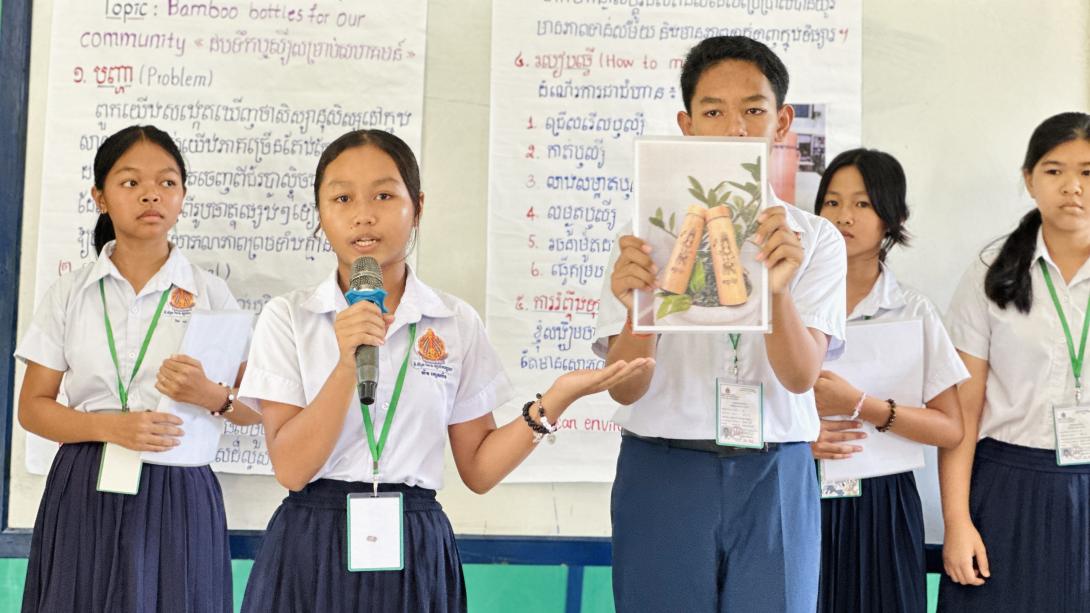 A group of school students in white short-sleeved shirts and dark skirts or trousers stand in front of a white board giving a presentation. A young woman in the front left holds a microphone and speaks, gesturing with her left hand toward a classmate beside her. The young man holds up a printed photograph showing two decorated bamboo bottles nestled in green leaves. In the background, posters with text in English and Khmer script are visible, with one heading reading "Topic: Bamboo bottles for our community