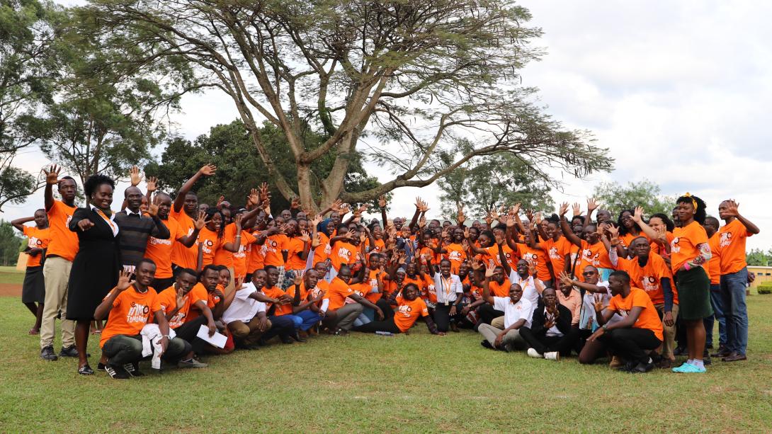 A wide shot of a large group of people posing together on a grassy lawn in front of a sprawling, leafy tree under an overcast sky. The majority of the group is wearing matching bright orange t-shirts with a graphic logo on the front, while a few individuals are in business or white shirts. Many people in the crowd are smiling and waving their hands at the camera. The individuals are arranged in rows, with those in the front kneeling or sitting on the grass, and those in the back standing.