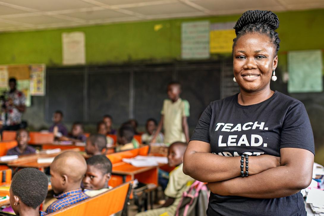 A smiling Black woman with her arms crossed wears a black T-shirt that reads "TEACH. LEAD." while standing in the foreground of a classroom filled with seated students.
