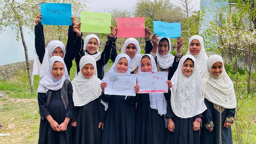 A group of young girls wearing white hijabs stand outdoors, holding up colorful signs that read "Happy 10th Anniversary Of Teach For Afghanistan."