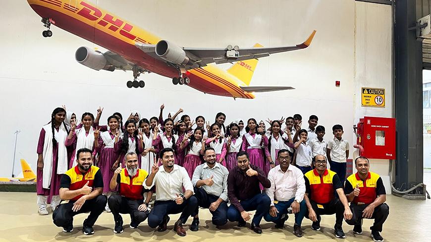 A group of school children in uniforms and several adults pose together indoors, with a large model of a DHL cargo plane mounted on the wall above them.