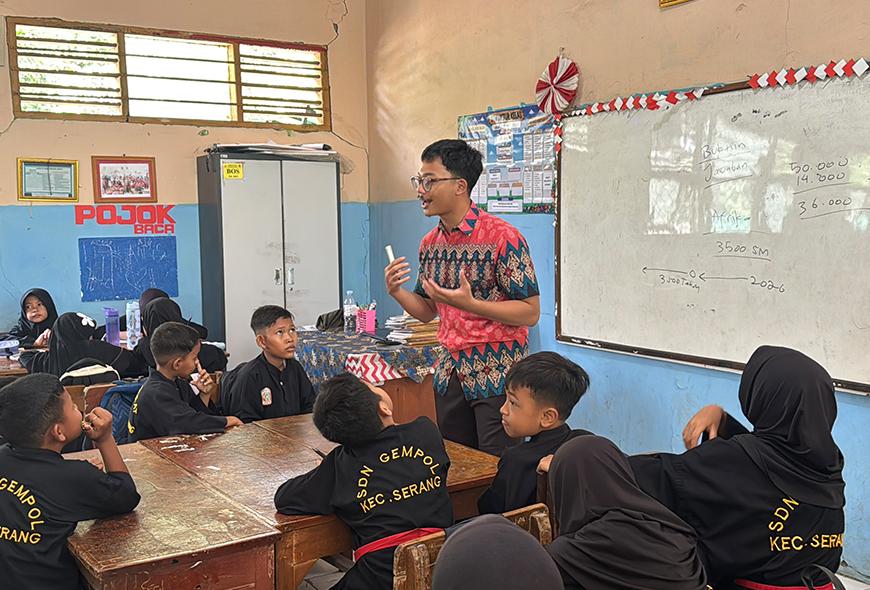 A teacher standing in front of a whiteboard, gesturing while presenting a lesson to students seated around tables in a classroom